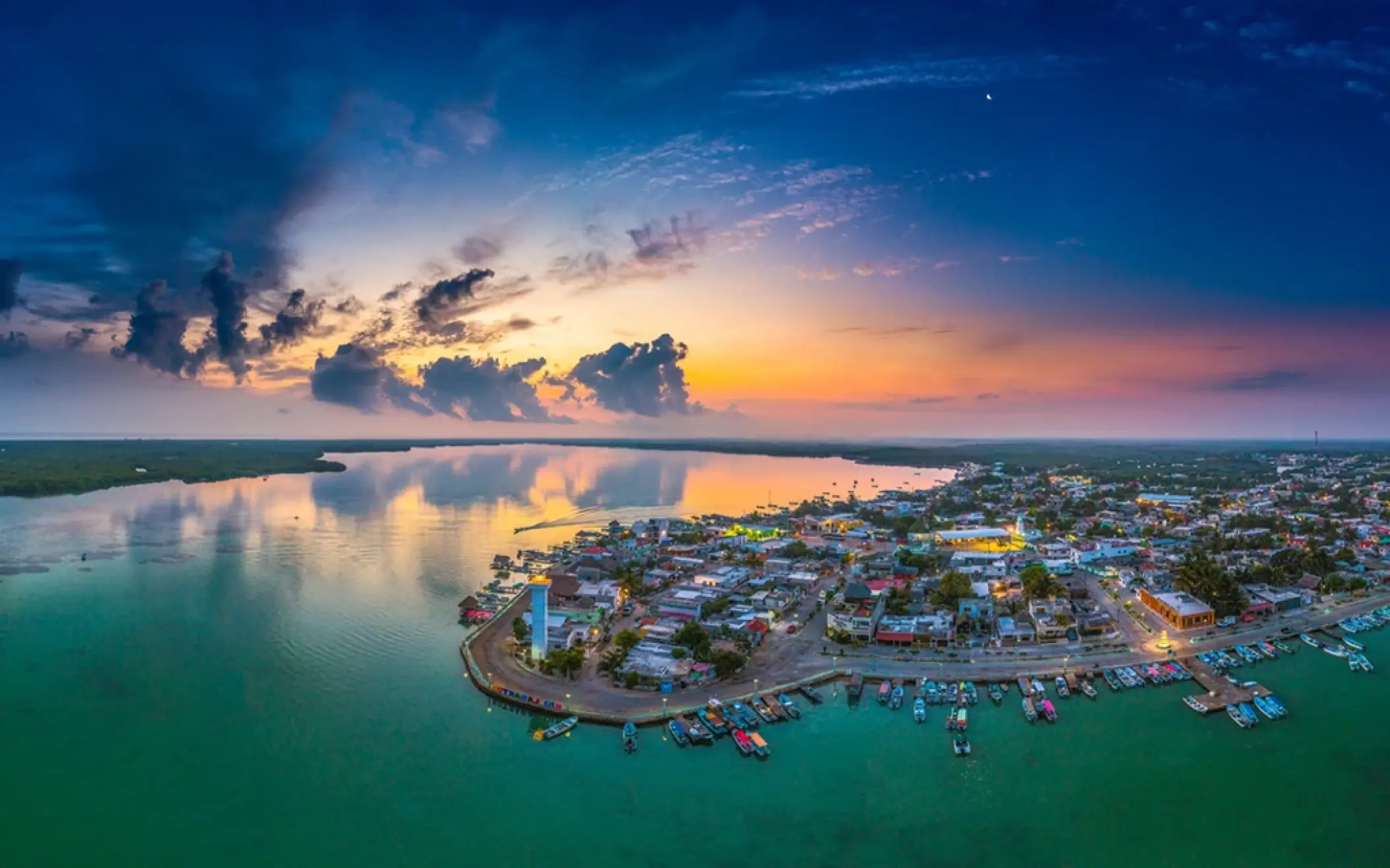 A stunning panoramic sunset view of the Rio Lagartos fishing village, with colorful boats docked along the calm, expansive lagoon.