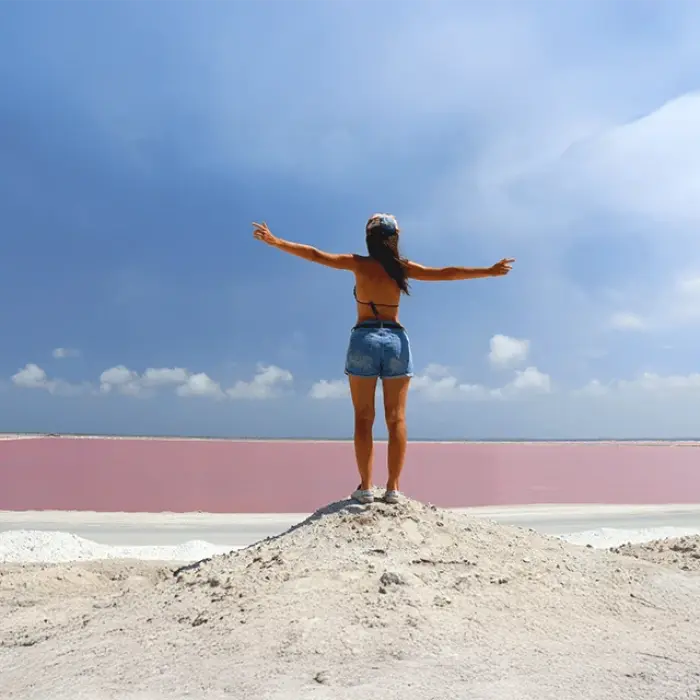 A woman stands with arms outstretched on a salt mound, overlooking the stunning pink lakes of Las Coloradas on a sunny day in Mexico.