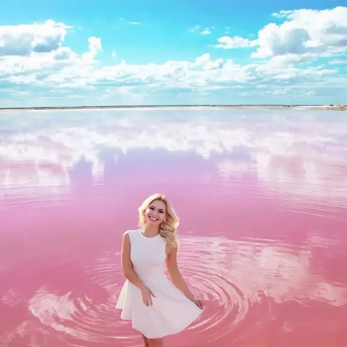 A smiling woman in a white dress wades in the magical pink water of Las Coloradas, with a beautiful sky reflecting on the lake.