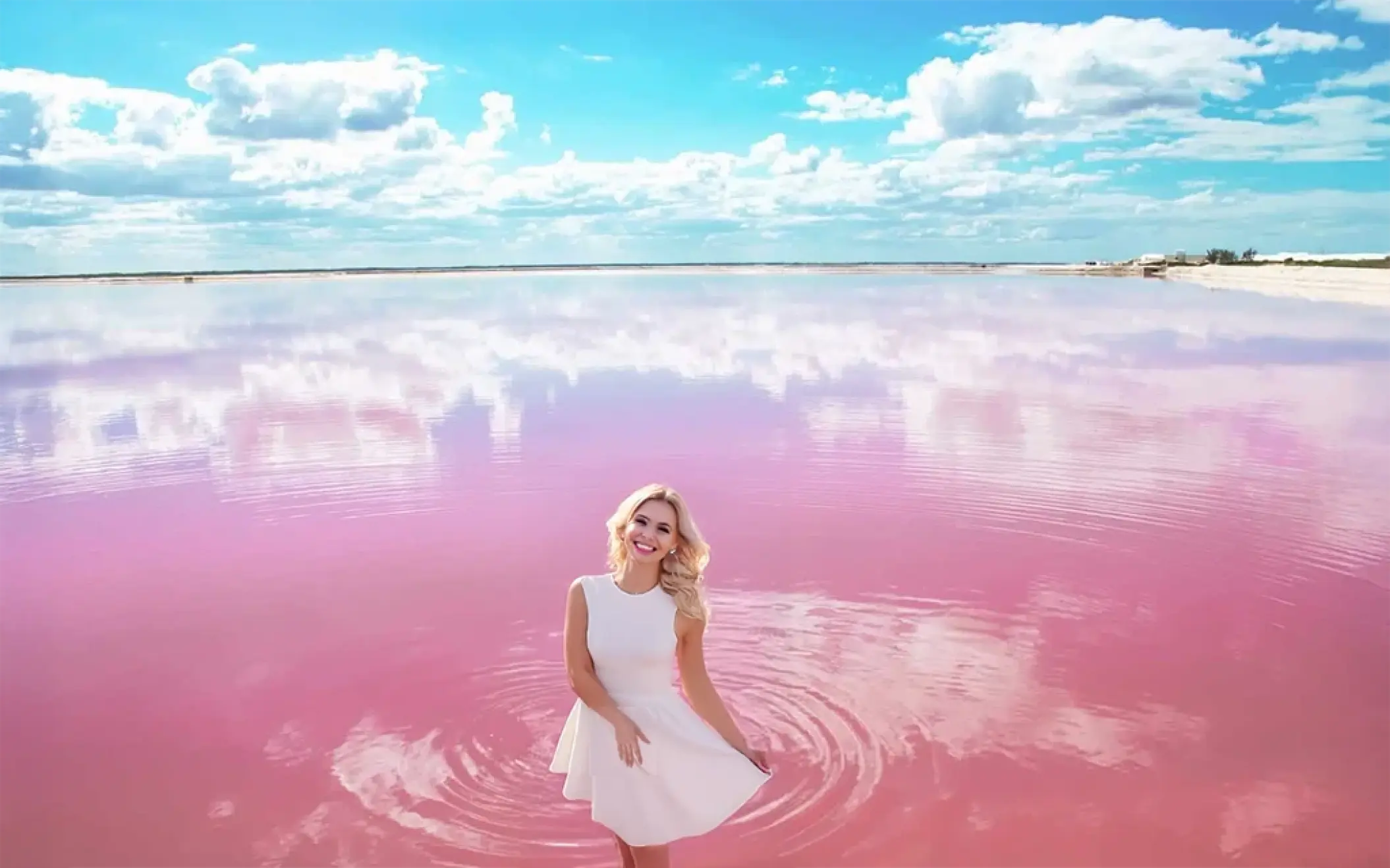 A smiling woman in a white dress wades in the magical pink water of Las Coloradas, with a beautiful sky reflecting on the lake.