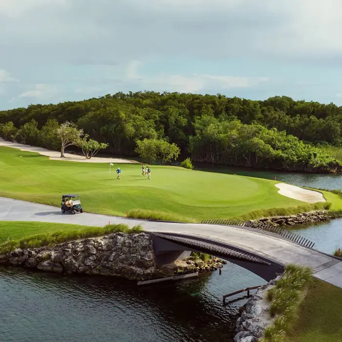 A golf cart crosses a beautiful stone bridge over a clear lagoon, heading towards the championship greens at El Camaleón Mayakoba.