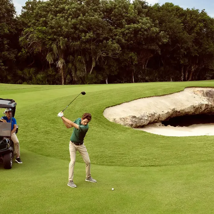 A golfer in a green shirt takes a powerful swing right next to the famous natural cenote cave bunker on the fairways of El Camaleón.