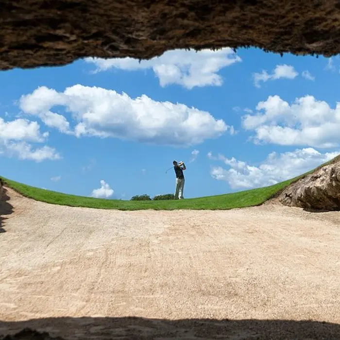A golfer teeing off from a unique cave bunker opening up to a bright blue sky at the famous El Camaleón golf course.