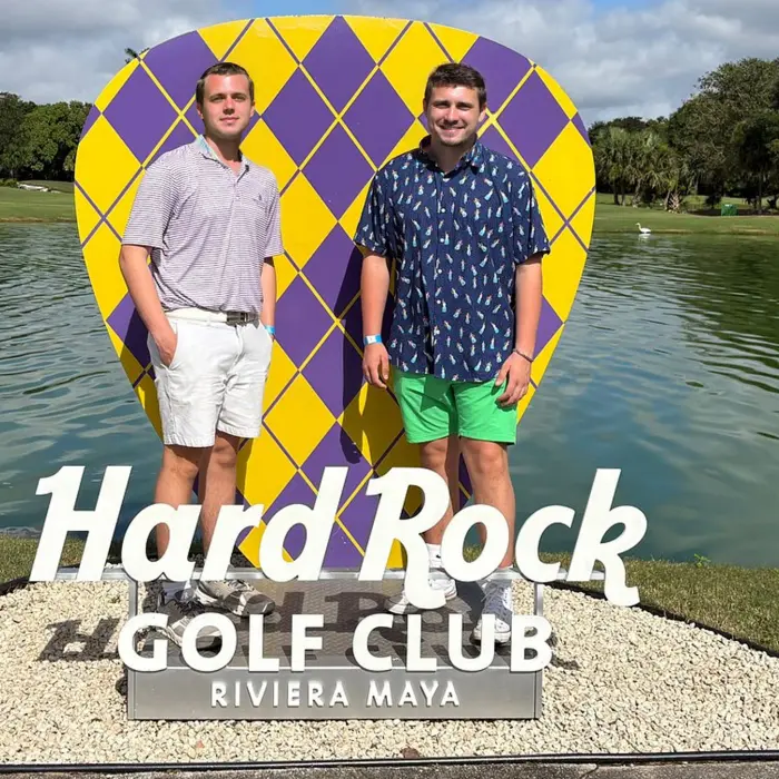 Two smiling golfers pose in front of a giant Hard Rock Golf Club guitar pick sign by a scenic lake in Riviera Maya.