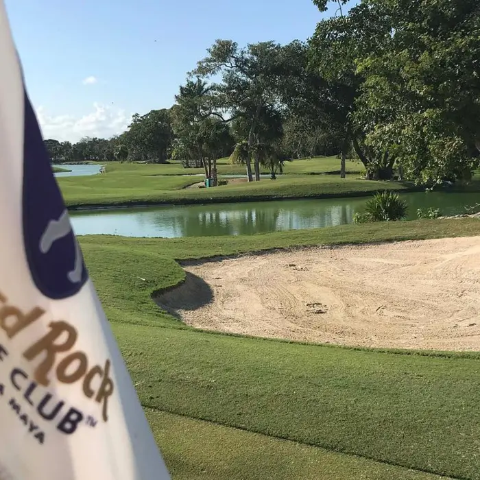 A close-up of a white Hard Rock Golf Club Riviera Maya flag fluttering on the green near a sand bunker and lake.