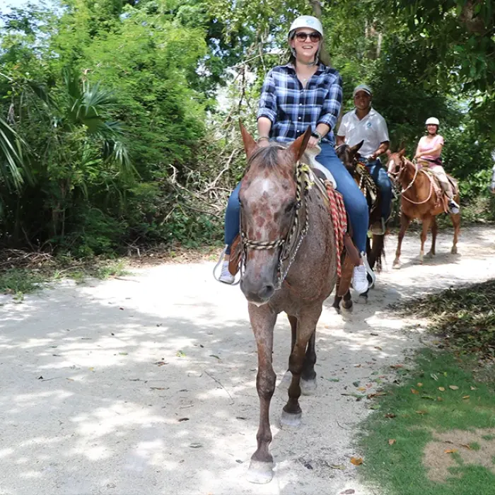 Horseback riding tour Playa del Carmen: Group of people wearing helmets riding horses on a sandy trail through a lush green forest