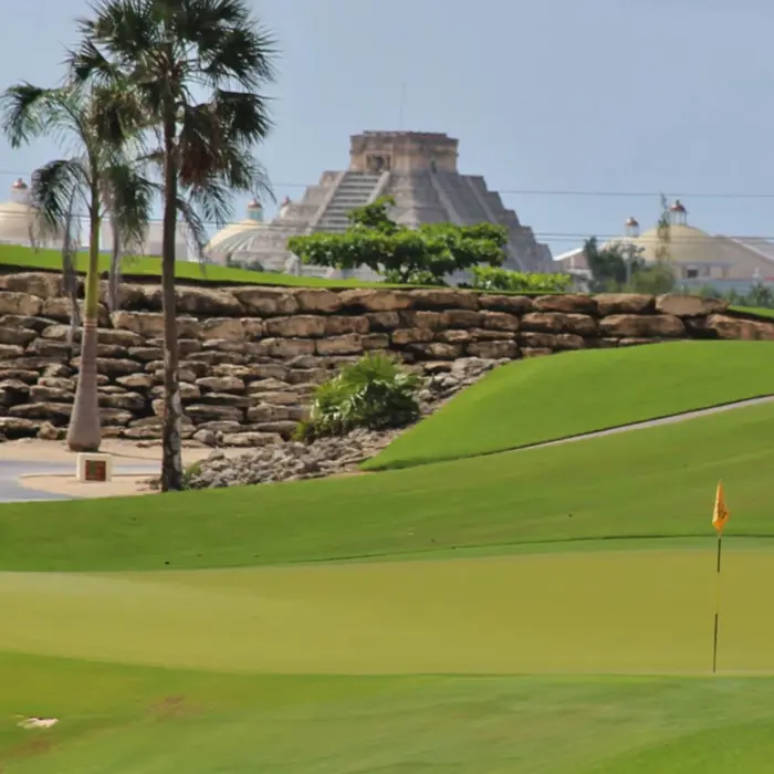 A yellow golf flag on a smooth green with a decorative rock wall and Mayan-inspired resort architecture in the distance.