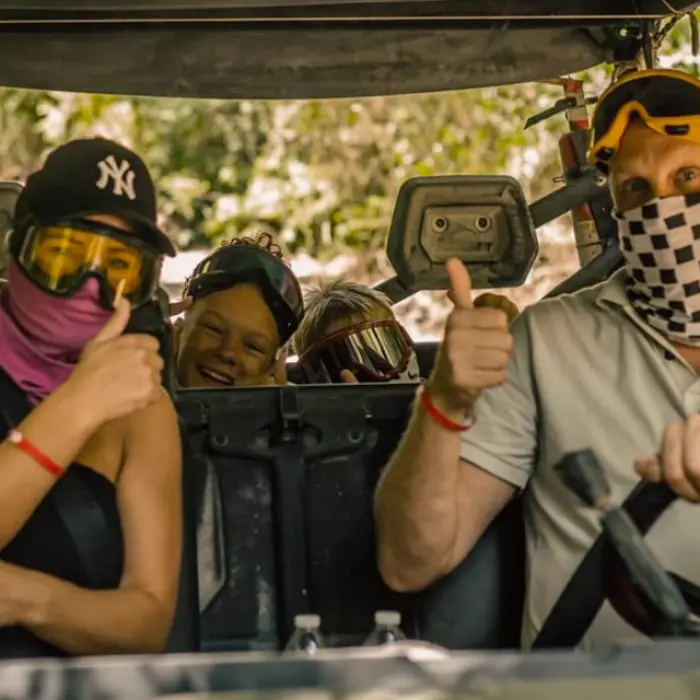 A group of adventure seekers inside an off-road buggy during a jungle tour, wearing masks, sunglasses, and protective gear while giving thumbs-up signs