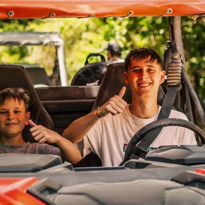 Two young adventurers smiling and giving thumbs-up while seated inside an orange off-road buggy during a jungle tour