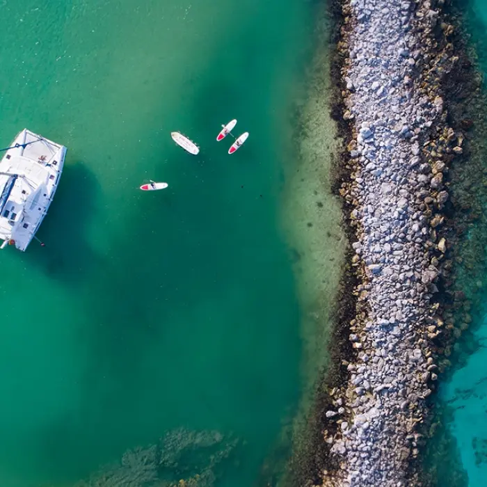 Aerial view of a catamaran near a rocky breakwater, with paddleboarders floating nearby