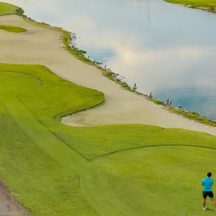 An elevated, scenic view of a couple enjoying a round of golf on a pristine green bordered by a sweeping sand bunker and a peaceful lake at Playa Mujeres Golf Club