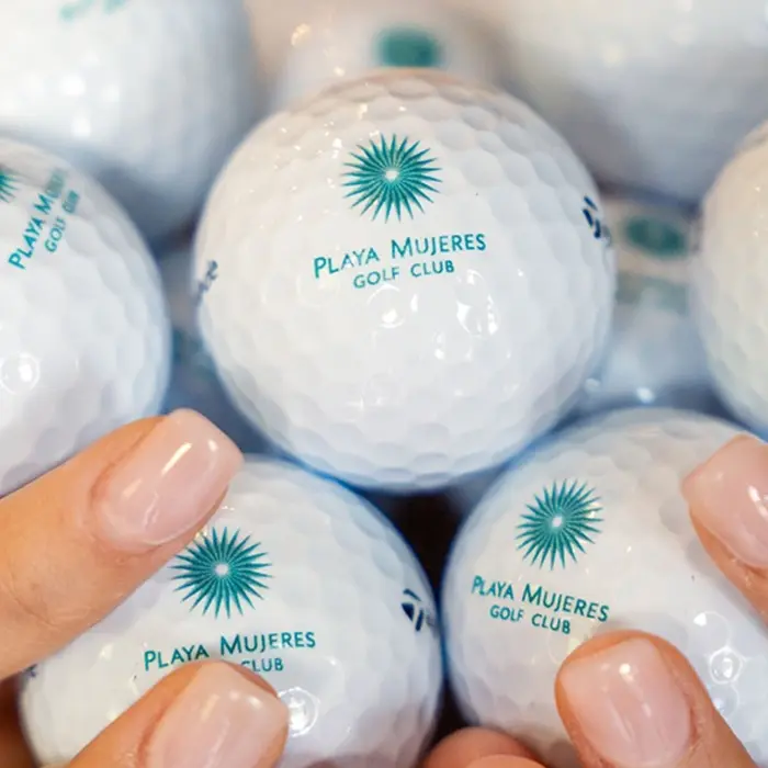 A close-up lifestyle shot of a golfer's hands holding a collection of premium TaylorMade golf balls featuring the elegant Playa Mujeres Golf Club logo