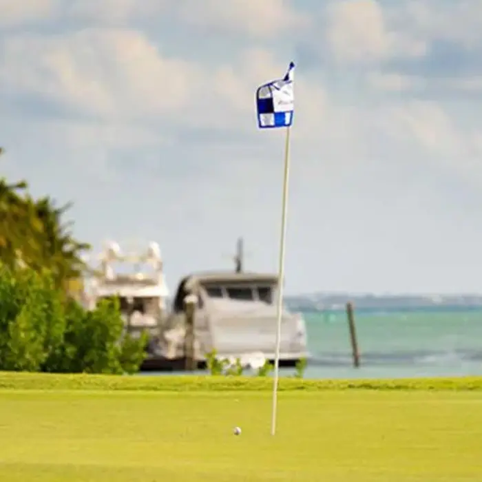 A checkered golf flag flutters in the ocean breeze on a pristine green with boats in the background at Puerto Cancún