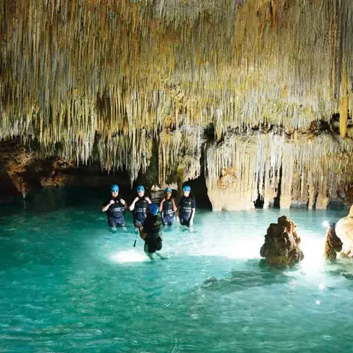 Rio Secreto cave exploration: Group of people wading through clear turquoise water in a cave filled with stalactites
