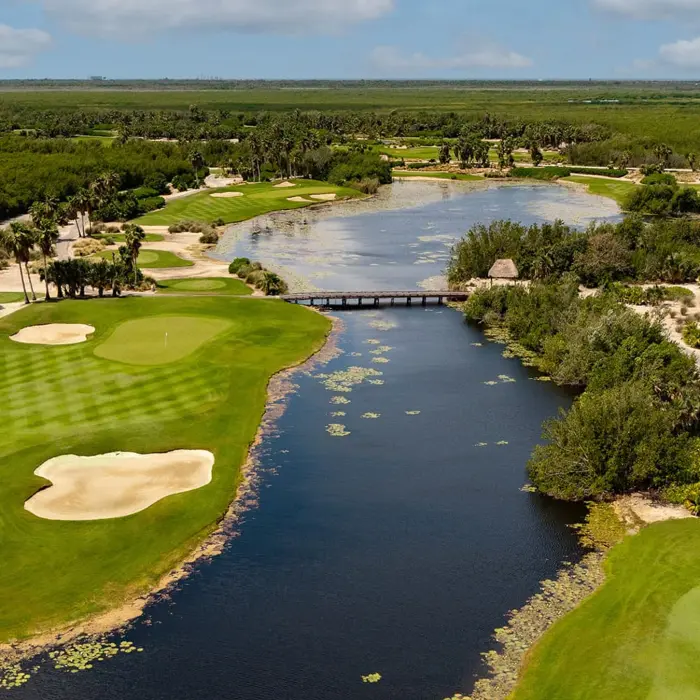 An expansive aerial view of the Jack Nicklaus Signature course at Riviera Cancún Golf Club, featuring smooth green fairways separated by a wide, scenic lake and a wooden bridge