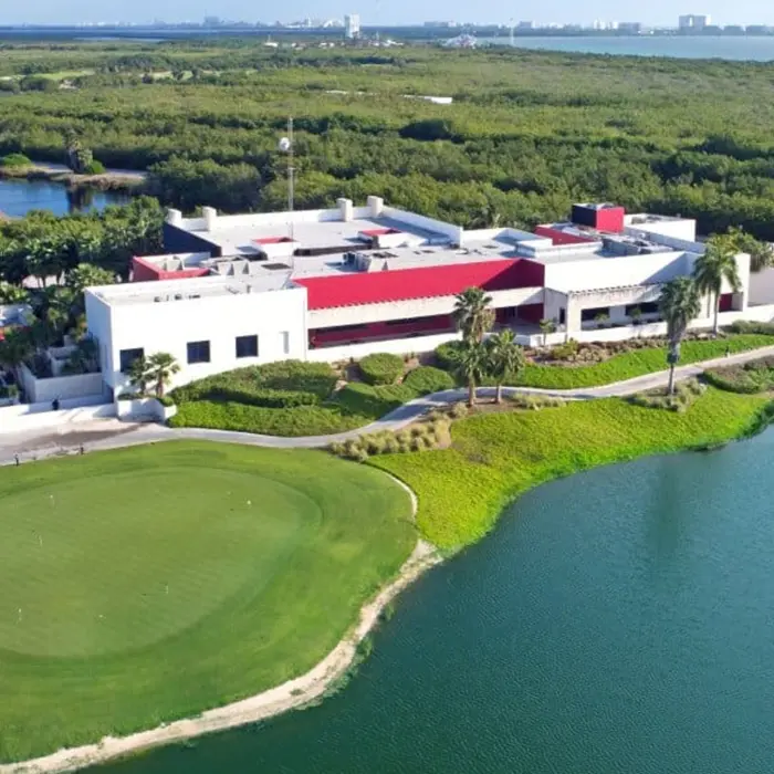 An aerial perspective of the modern luxury clubhouse at Riviera Cancún Golf Club, showing its striking red and white architecture adjacent to the practice greens and a large lake