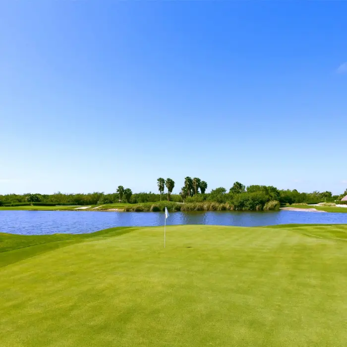 A perfectly maintained, elevated green with a white flag at Riviera Cancún Golf Club, situated beside a peaceful lake reflecting the clear blue sky