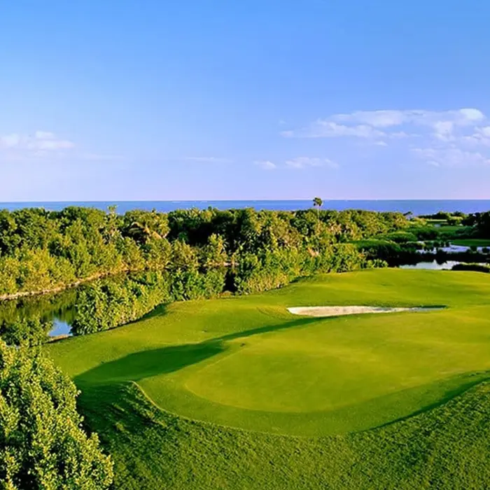 A beautiful elevated view of a golf green at Riviera Cancún Golf Club, bordered by sand bunkers and surrounded by lush tropical jungle and tranquil lakes under a blue sky