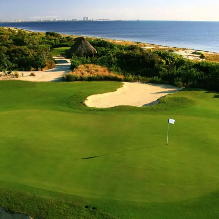 A pristine golf green positioned right along the serene coastline, with gentle waves and the Caribbean Sea visible in the background at Riviera Cancún Golf Club