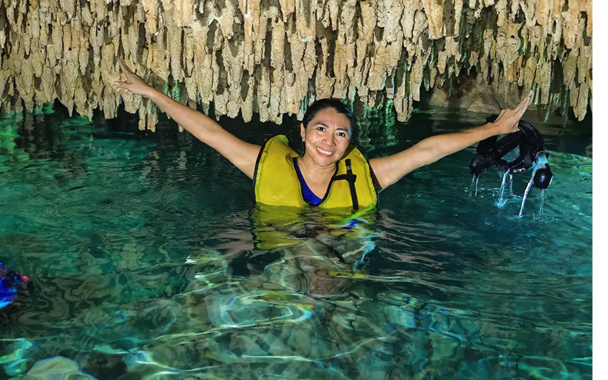 Smiling woman swimming in a cenote cave, wearing a life jacket and holding goggles