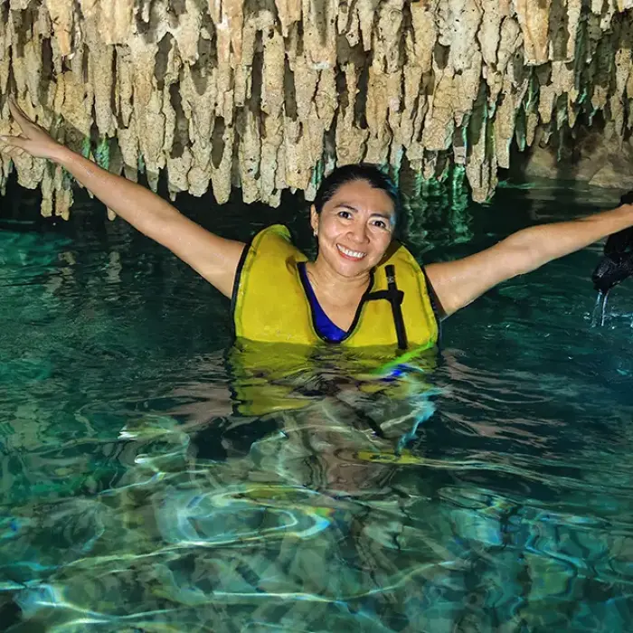 Smiling woman swimming in a cenote cave, wearing a life jacket and holding goggles