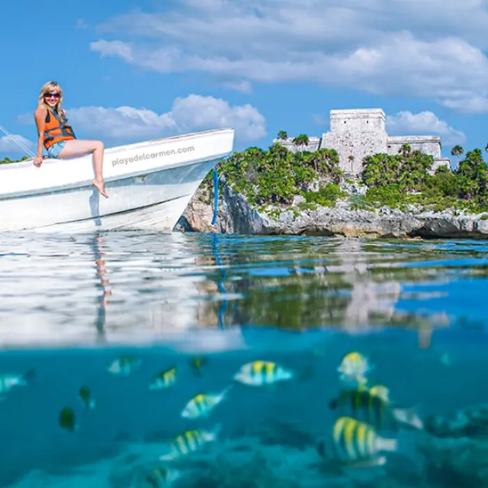 A woman relaxing on a boat near the coast, with the Tulum ruins in the background and tropical fish swimming below