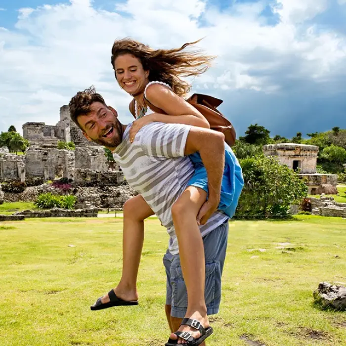 Man giving woman a piggyback ride with big smiles among the ruins of Tulum