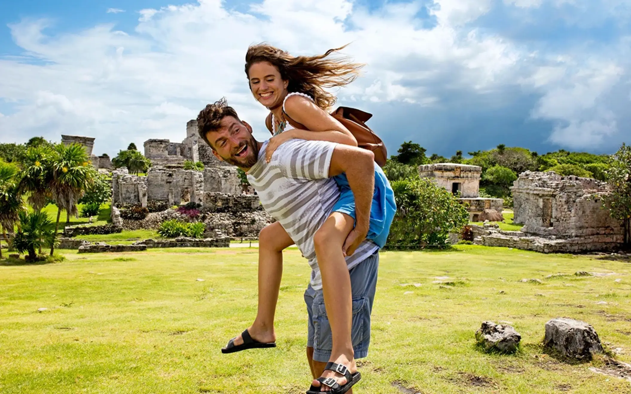 Man giving woman a piggyback ride with big smiles among the ruins of Tulum