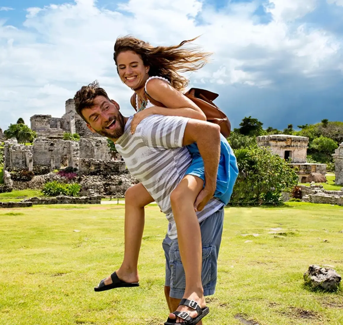 Man giving woman a piggyback ride with big smiles among the ruins of Tulum