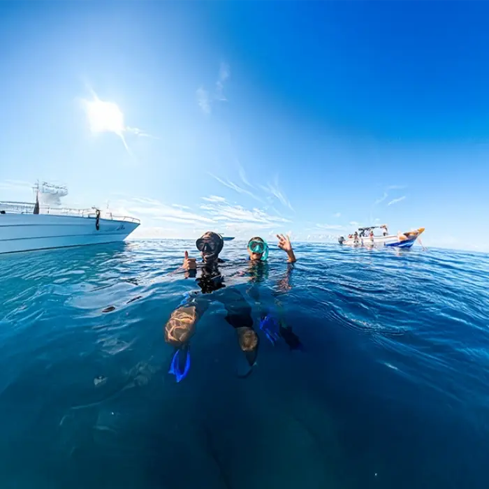 Two happy snorkelers give a thumbs-up in the beautiful blue ocean after their swim, with our comfortable and modern tour boat behind them.