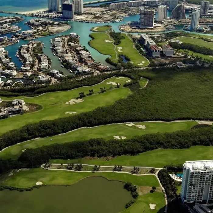 Expansive aerial view of the championship golf course fairways winding through lush mangroves with luxury high-rises and the Caribbean Sea in the distance