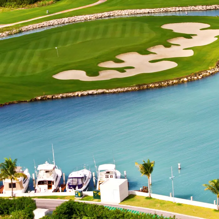 An aerial perspective of a stunning island golf green surrounded by crystal clear blue water with luxury yachts docked in the foreground.