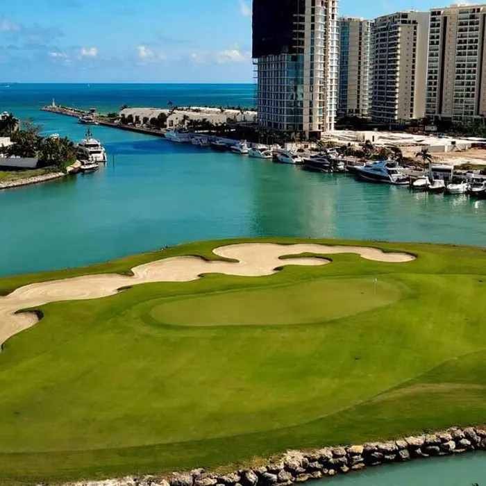 An aerial top-down view of a vibrant golf green and a uniquely shaped sand bunker bordering the bright turquoise waters at Puerto Cancún Golf Club