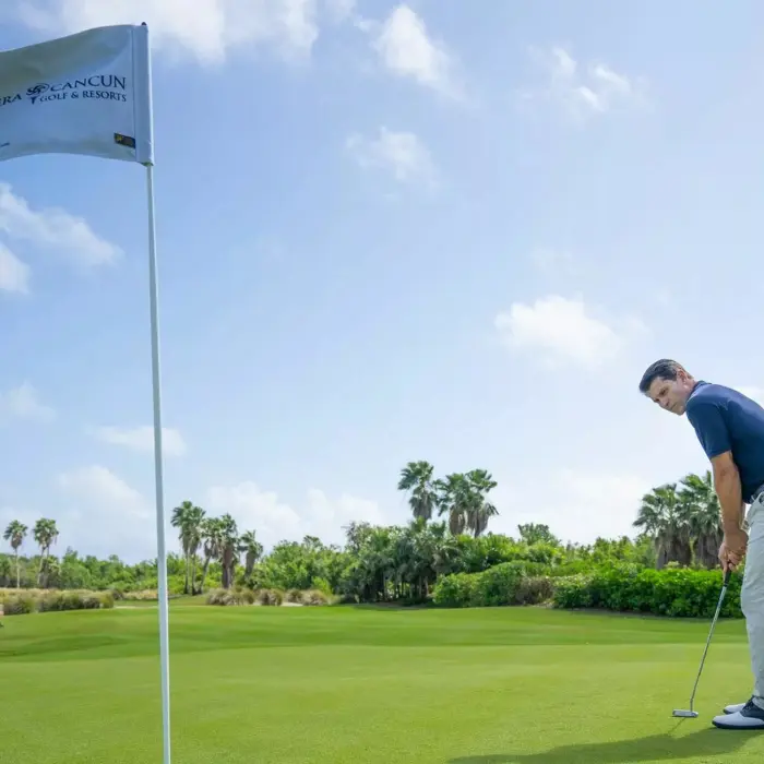 A focused golfer carefully lining up a putt on a perfectly smooth championship green under a clear blue sky in the Riviera Maya.