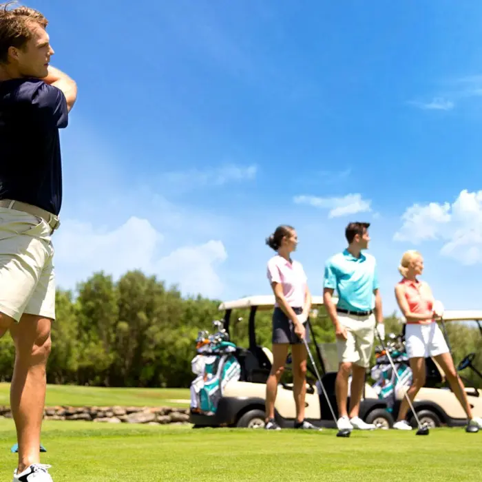 A dynamic lifestyle shot of a golfer completing his swing on the tee box, while his friends watch near their golf carts under a bright blue sky.