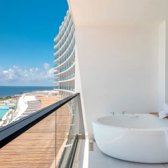 A large white soaking tub on a balcony overlooking the ocean and pool area.