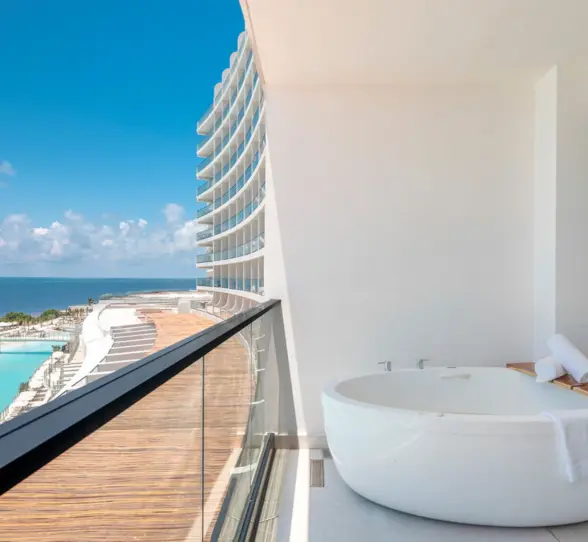 A large white soaking tub on a balcony overlooking the ocean and pool area.