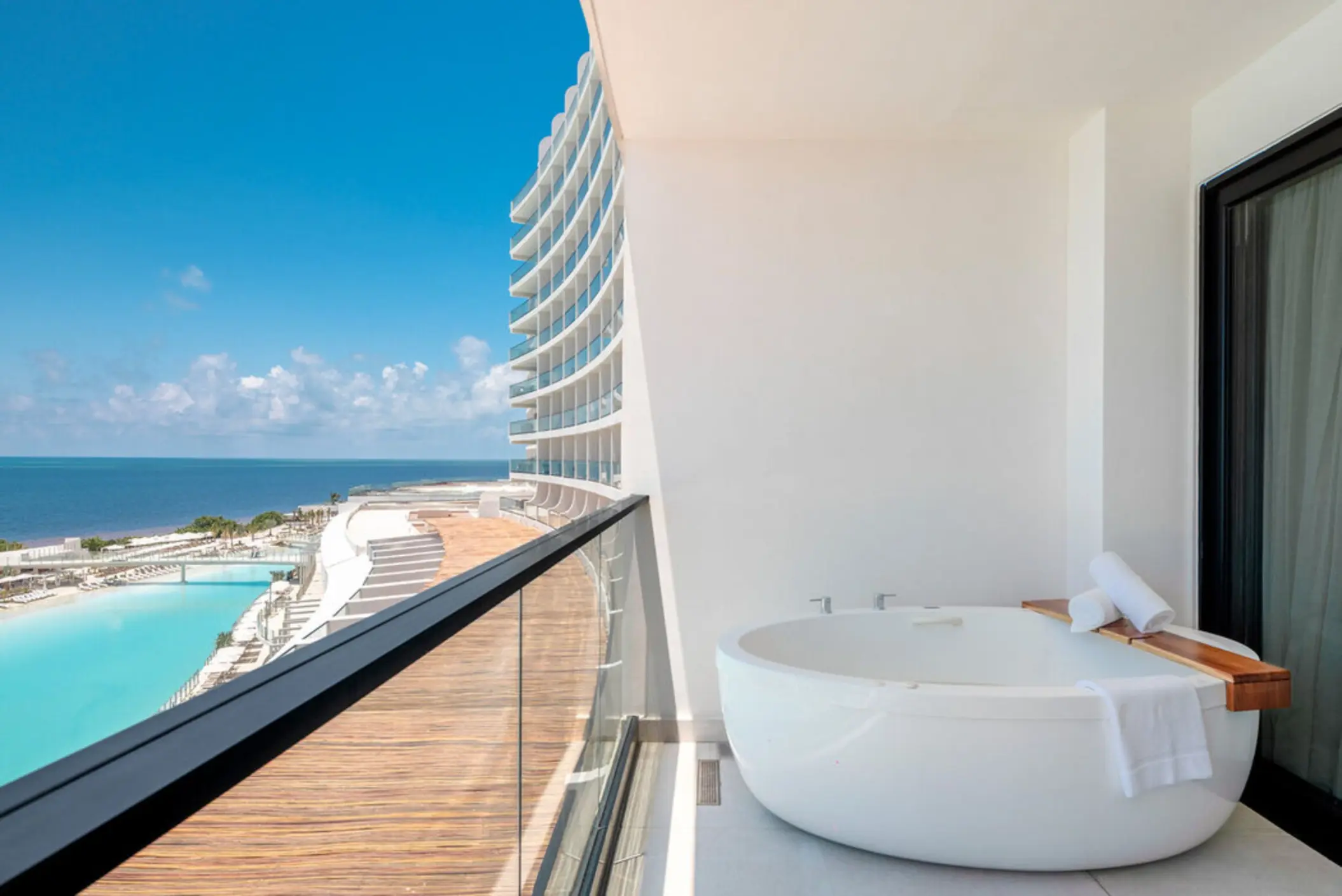 A large white soaking tub on a balcony overlooking the ocean and pool area.