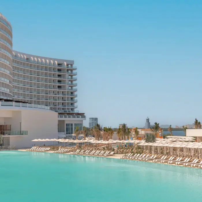 A wide, turquoise-colored pool with rows of lounge chairs, next to a modern resort building.