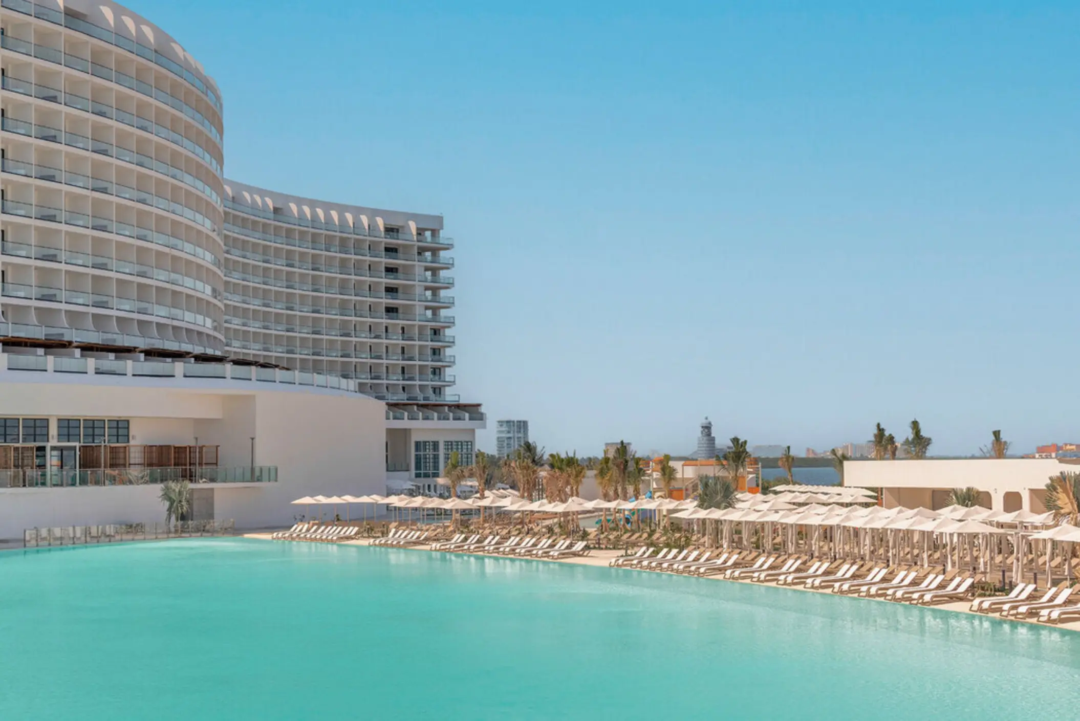 A wide, turquoise-colored pool with rows of lounge chairs, next to a modern resort building.