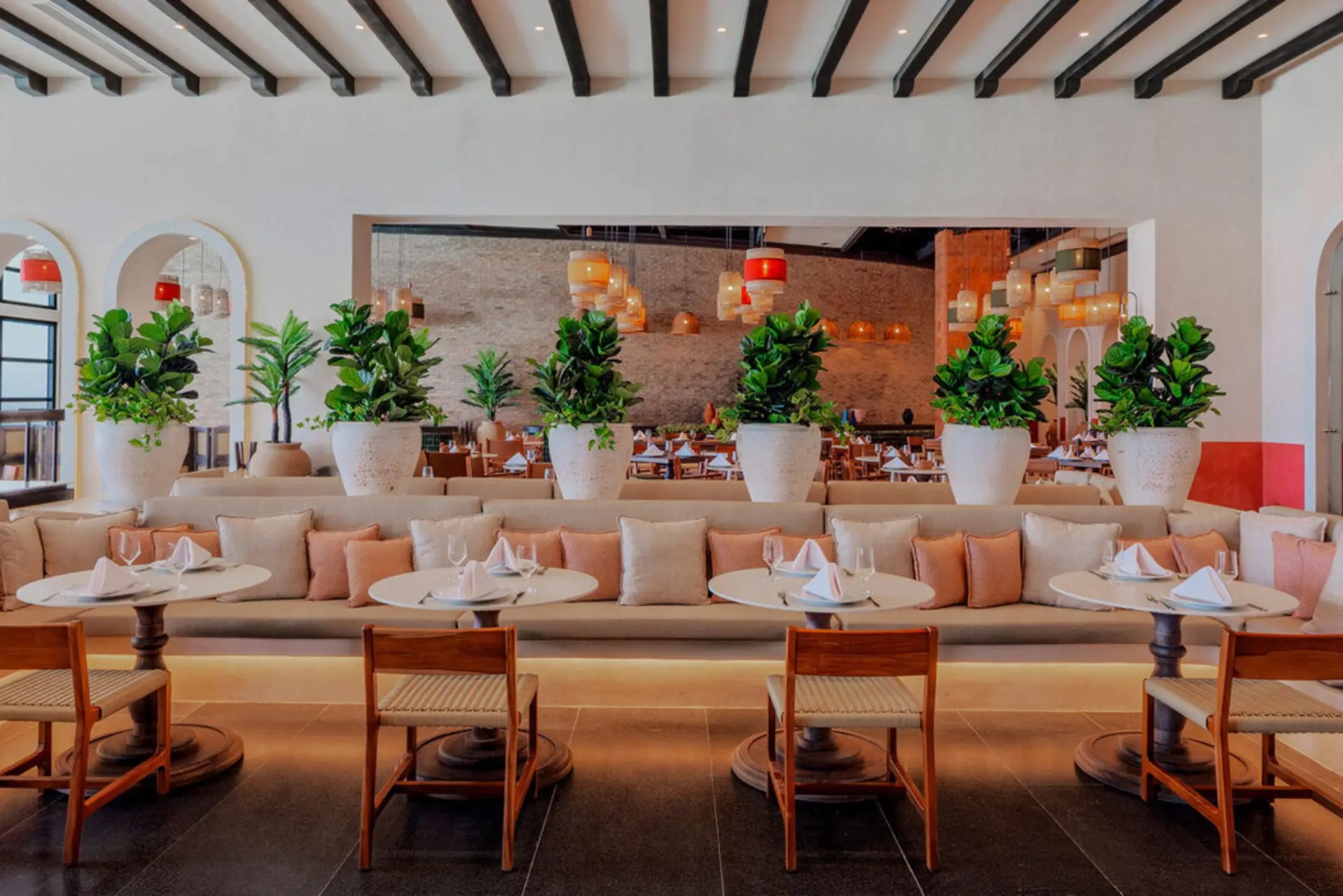 The interior of a contemporary restaurant with long banquettes, round tables, and potted plants.