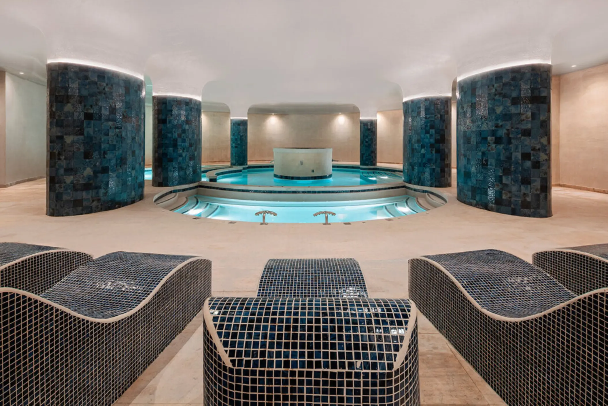 An indoor spa area with a round pool, blue tiled walls, and chaise lounges.