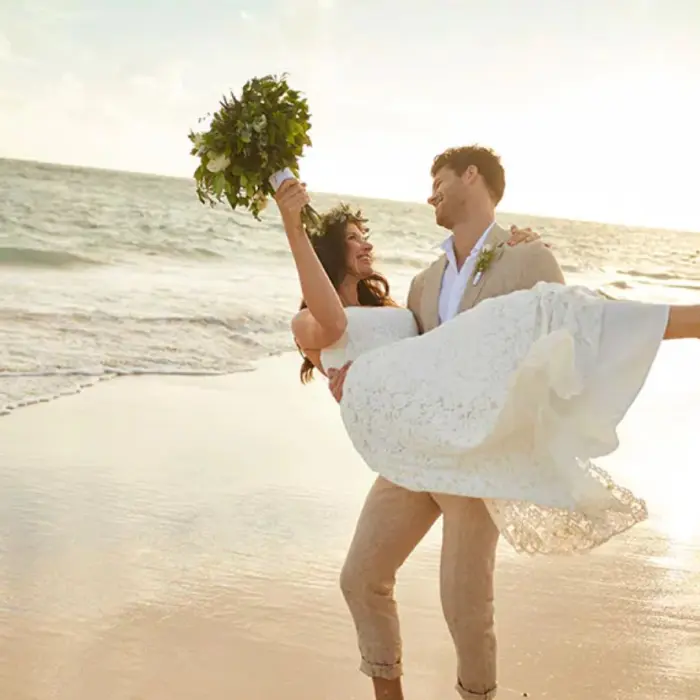 Bride held by the groom on a sandy beach with the ocean and sunset in the background, celebrating at Bahia Principe Grand Coba.