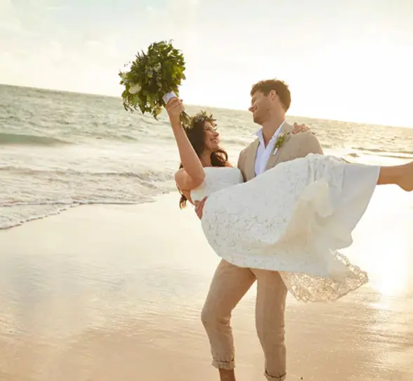 Bride held by the groom on a sandy beach with the ocean and sunset in the background, celebrating at Bahia Principe Grand Coba.