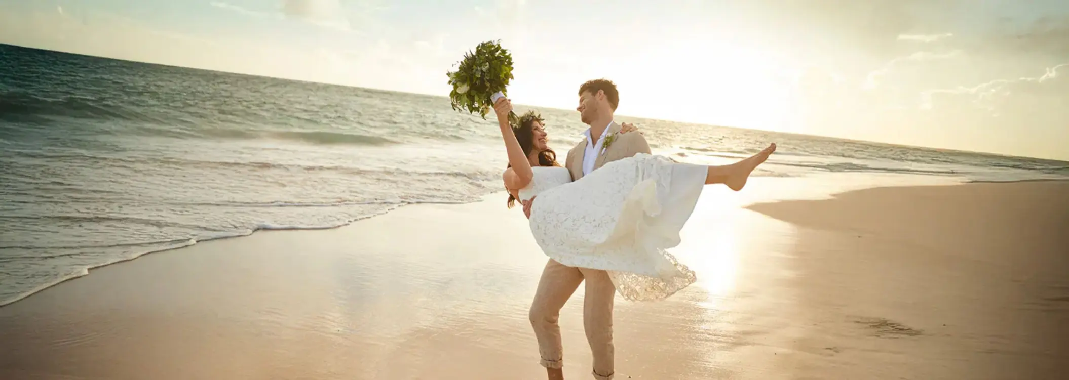 Bride held by the groom on a sandy beach with the ocean and sunset in the background, celebrating at Bahia Principe Grand Coba.