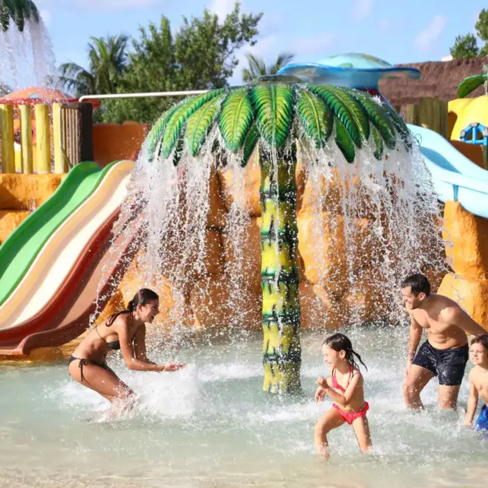 Family enjoying a splash pad with a large mushroom water feature and colorful slides at Bahia Principe Grand Coba.