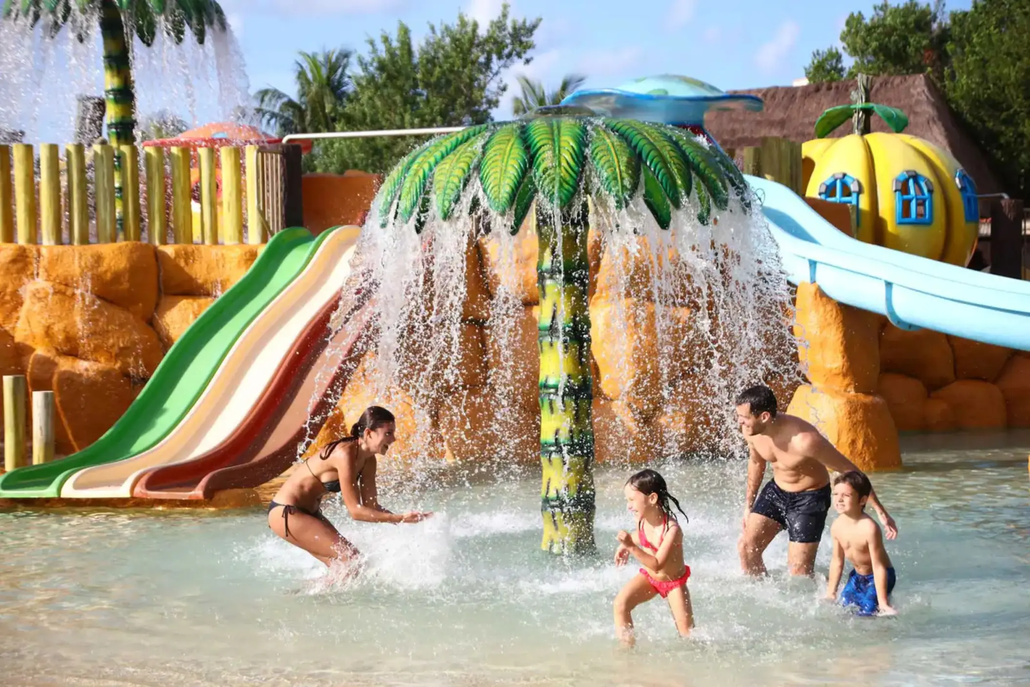 Family enjoying a splash pad with a large mushroom water feature and colorful slides at Bahia Principe Grand Coba.