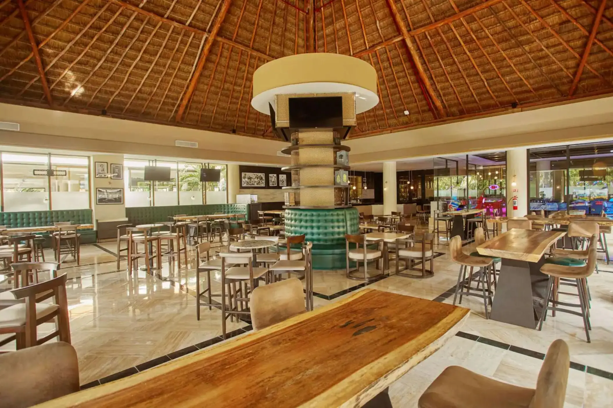 Interior of a resort restaurant with various seating arrangements and a high thatched roof at Bahia Principe Grand Coba.