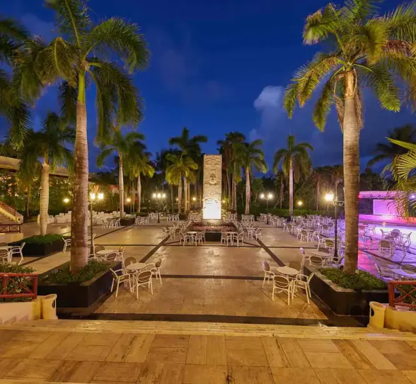 Outdoor dining area at night, illuminated by lights, with palm trees and tables set up at Bahia Principe Grand Coba.