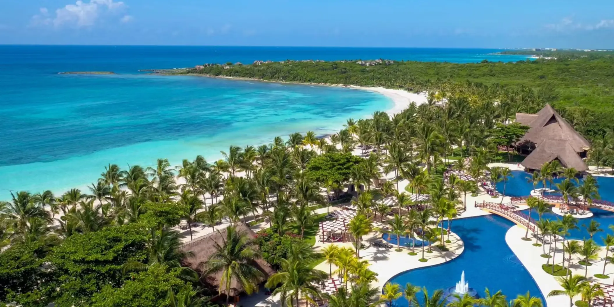 aerial view of Barceló Maya Beach resort, showcasing large pools, lush palm trees, and the expansive white sand beach bordering the turquoise ocean.